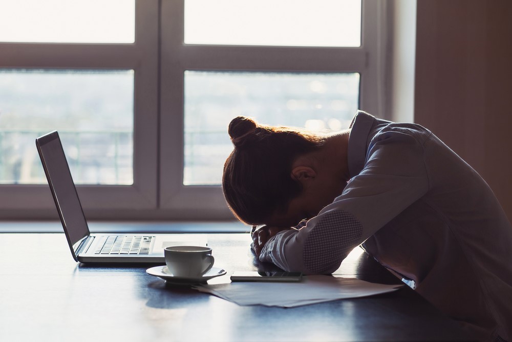 A woman sitting at the table resting her head on her arms with a laptop and papers in front of her. 