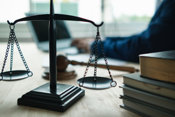 Close-up of a balance scale on a desk with a gavel and stacked law books, while a blurred person works on a laptop in the background.