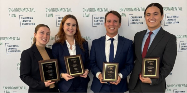 Four people in business attire stand side by side. each smiling and holding a framed award plaque.